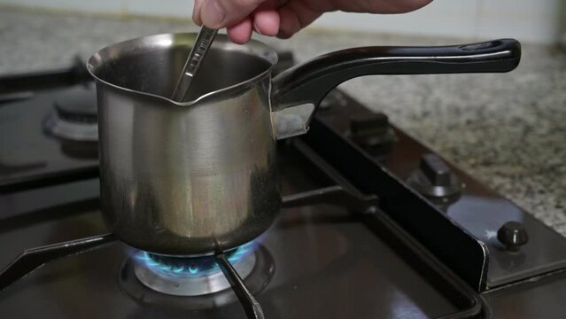 A man's hand stirs with a spoon inside a coffee warmer pan. The coffee warmer pan is positioned over an awaiting flame, slowly extinguishing.
