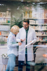 View through the window of the pharmacy of a healthcare worker helping a senior woman.