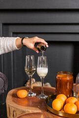 The woman's hand pours champagne from a bottle into glass, tangerines on a table against blurred black background. New year celebration party.Winter holidays celebration