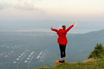 Wide shot of back of woman wear red coat and stand with spread her arms near cliff on the mountain and look forward to beautiful view with warm light of sun rising.