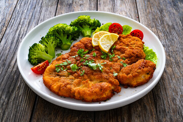 Crispy breaded fried chicken cutlet with boiled broccoli and fresh vegetables on wooden table
