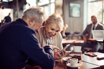 Senior man and woman laughing pointing at smartphone in cafe