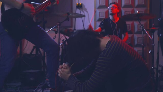 The lead singer of a rock band, kneeling on the floor, belts out a song with emotion as the electric guitarist and drummer accompany him. The band is rehearsing for a performance in a rehearsal room.
