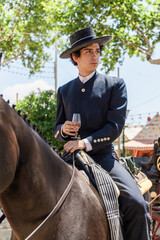 Stylish young man in hat sitting on horse with wineglass in hand in countryside