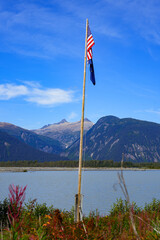 United States and Alaskan flags hanging at the top of a wooden flagpole on the shores of a melt water lake connected to the Taku Inlet in the mountains north of the Alaskan capital city Juneau