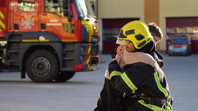 Brave firefighter in uniform hugs a little boy against a fire engine