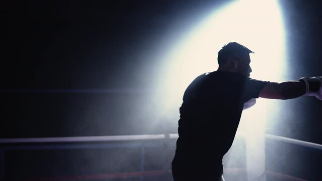 Young fighter punching the air inside boxing ring with dramatic lighting spotlight and haze. Sportsman athlete training before match