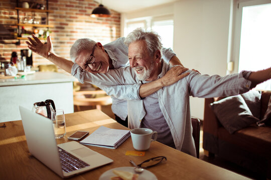 Happy senior man with outstretched arms looking at laptop with wife in home