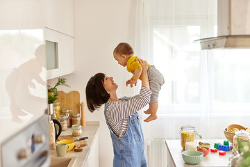 Mother lifting up her baby boy in the air in the kitchen - obrazy, fototapety, plakaty