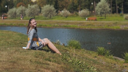 Naklejka na ściany i meble A girl sits in a meadow on a summer day by the river.