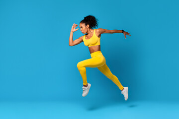 Fototapeta na wymiar African Lady In Fitwear Exercising Jumping Over Blue Studio Background