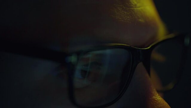 The eyes of a businessman in the spotlight, working at night, intently study information and analyze data using a portable laptop. Reflections flicker in his glasses.