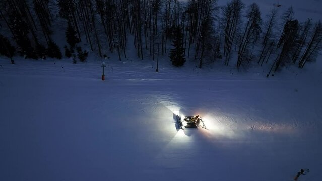 Aerial view of working snowcat on the snowy alpine ski slope at winter night in Kranjska Gora, Slovenia. Top drone view of snowcat grooming snow, ski resort, lights at dusk. Snowplough machine