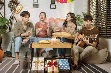 Group of Asian male and female friends sitting at a party at a friend's house, playing guitar and singing, drinking wine, having fun together, living room at a friend's house.