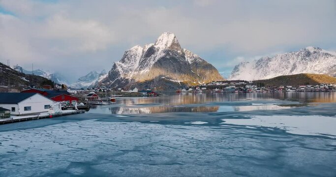Aerial view of snowy rocks, islands with rorbuer, frozen sea coast, mountains, road, sky with clouds at sunset in winter. Beautiful top drone view of rorbu in Reine village, Lofoten islands, Norway