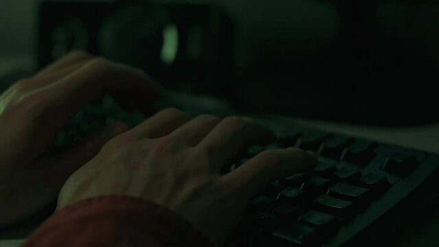 Under dim lighting man is busy working at his desk with a laptop computer. His confident movements on the keyboard create code. An IT specialist works in conditions of remote work or distance learning