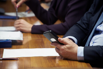 Man in a business suit uses a mobile phone - smartphone, sitting at a table during a meeting,...