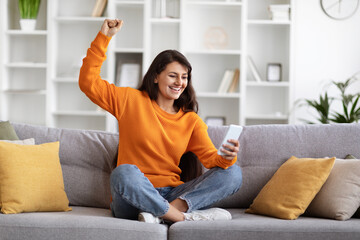 Excited young indian woman sitting on couch, holding smartphone