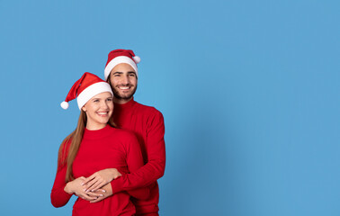 Obraz premium Happy young couple in Santa hats hugging on blue studio background