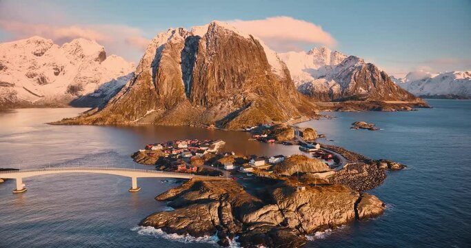 Aerial view of snowy rocks, islands with rorbuer, sea, bridge, mountains, road, sky with clouds at pink sunrise in winter. Beautiful top drone view of rorbu in Hamnoy village, Lofoten islands, Norway	