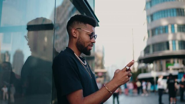 Cheerful Black young man, wearing glasses and adorned with necklaces, beams with laughter at phone screen, which display witty chat or delightful social media post, he stands on bustling street corner