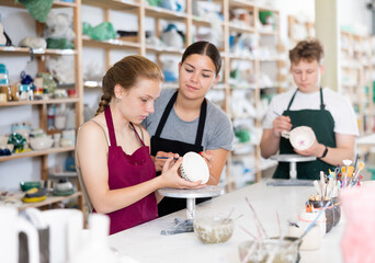 Young woman teacher showing teenage boy and teenage girl how to paint ceramic cup in workshop