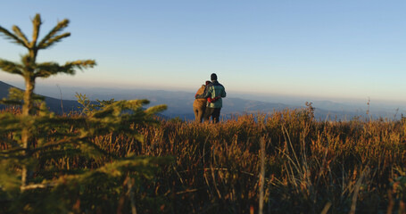 Multiethnic couple of tourist walks on top of hill in autumn. Caucasian woman and African American man hold hands, talk and look at sunset. Hikers enjoy view on their vacation in mountains.