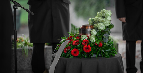 Metal urn or funeral container with ash of a deceased person at a memorial service. Undertakers seen in the back and a beautiful bouquet surrounding the urn. - obrazy, fototapety, plakaty
