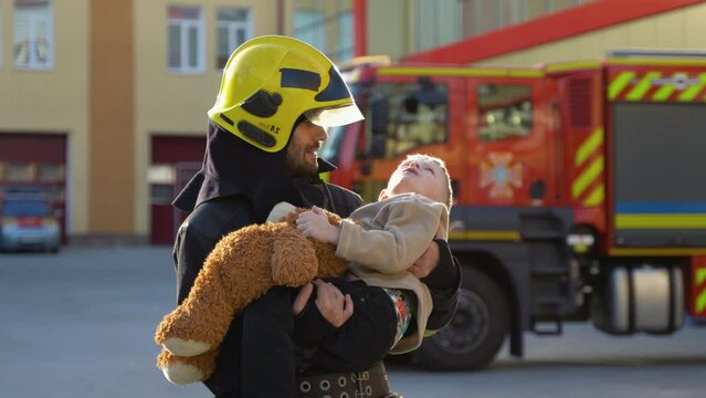 A firefighter take a little child boy to save him. Fireman with kid in his arms. Fire engine car on background