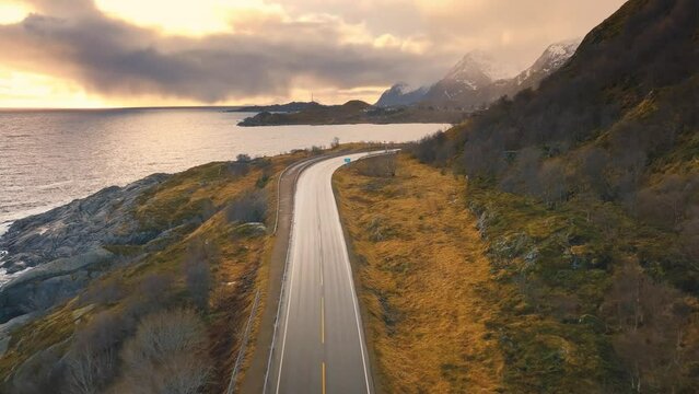 Aerial view of road, sea with waves, snowy mountains at sunset in winter. Lofoten Islands, Norway. Top view of beautiful road, orange grass, water, rocky beach, sky with low clouds, golden sunlight