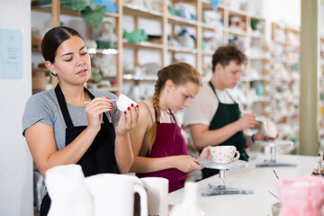 Fototapeta premium Young woman teacher in apron paints ceramic cup in ceramic workshop..