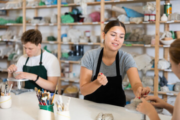 Fototapeta premium Young woman teacher shows students teenage girl and teenage boy how to sculpt ceramic product from raw clay in workshop