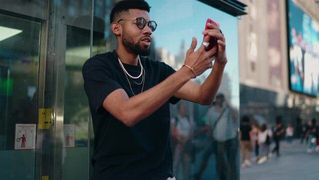 Fashionable Black man with glasses capturing selfie on smartphone in urban setting,express digital era connectivity and mobile technology. Millennial cellphone user having video call on frontal camera