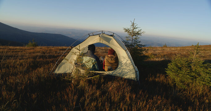 Multiethnic tourist couple sit in tent on top of mountain hill: They talk and drink tea. Two travelers stopped to rest during adventure vacation. Romantic hiker family admire the scenery. Back view. - Powered by Adobe