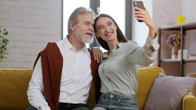 Young happy woman visiting her grandfather at home, making together selfie on smartphone, hugging and smiling to camera