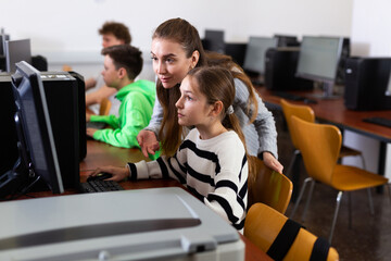 Positive female teacher helping concentrated teen schoolgirl studying in college library computer...