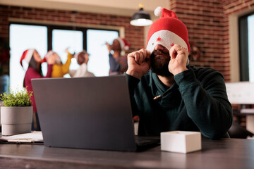 Frustrated tired employee putting santa hat on eyes while working overtime during christmas season...