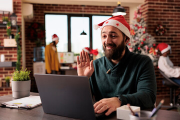 Smiling employee in santa hat waving hi while speaking at online corporate meeting using laptop....