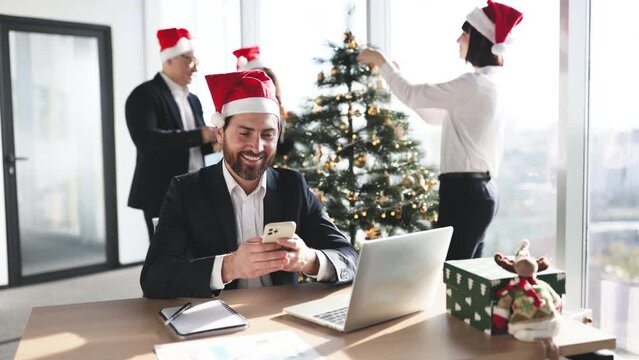 Businessman in Santa hat sitting at table and using modern smartphone in decorated festive office. Male freelancer taking break during working process while coworkers are decorating the Christmas tree