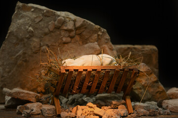 Manger with baby, hay and stones on wooden table against dark background - obrazy, fototapety, plakaty