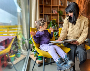 A grandmother reads a book to her granddaughter. A woman reads a book to a child, a girl, sitting in yellow chairs by the windo , family, joy, emotion