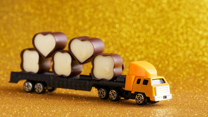 Toy truck with a trailer transports chocolates on a sparkling golden holiday background. Concept for delivering sweets for the holiday. Photo. Selective focus.