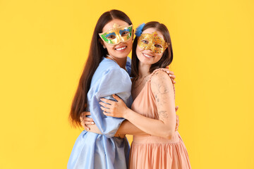 Beautiful young women in carnival masks hugging on yellow background