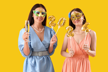 Beautiful young women with carnival masks, champagne and balloons in shape of figure 2024 on yellow background