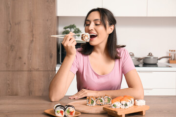 Happy young woman eating tasty sushi rolls in kitchen