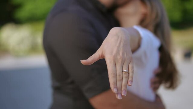 A woman, in the arms of a man, shows her hand with a wedding ring to the camera. Kissing couple out of focus