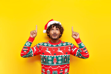 shocked young curly hindu man in new year clothes pointing with hands up on yellow isolated...