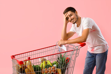 Tired young man with shopping cart and grocery list on pink background