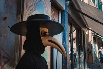 A man in a traditional plague mask stands on a spooky street in Venice, Italy, evoking the...
