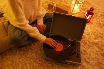 Woman in warm clothes with record player sitting on soft carpet in living room at evening